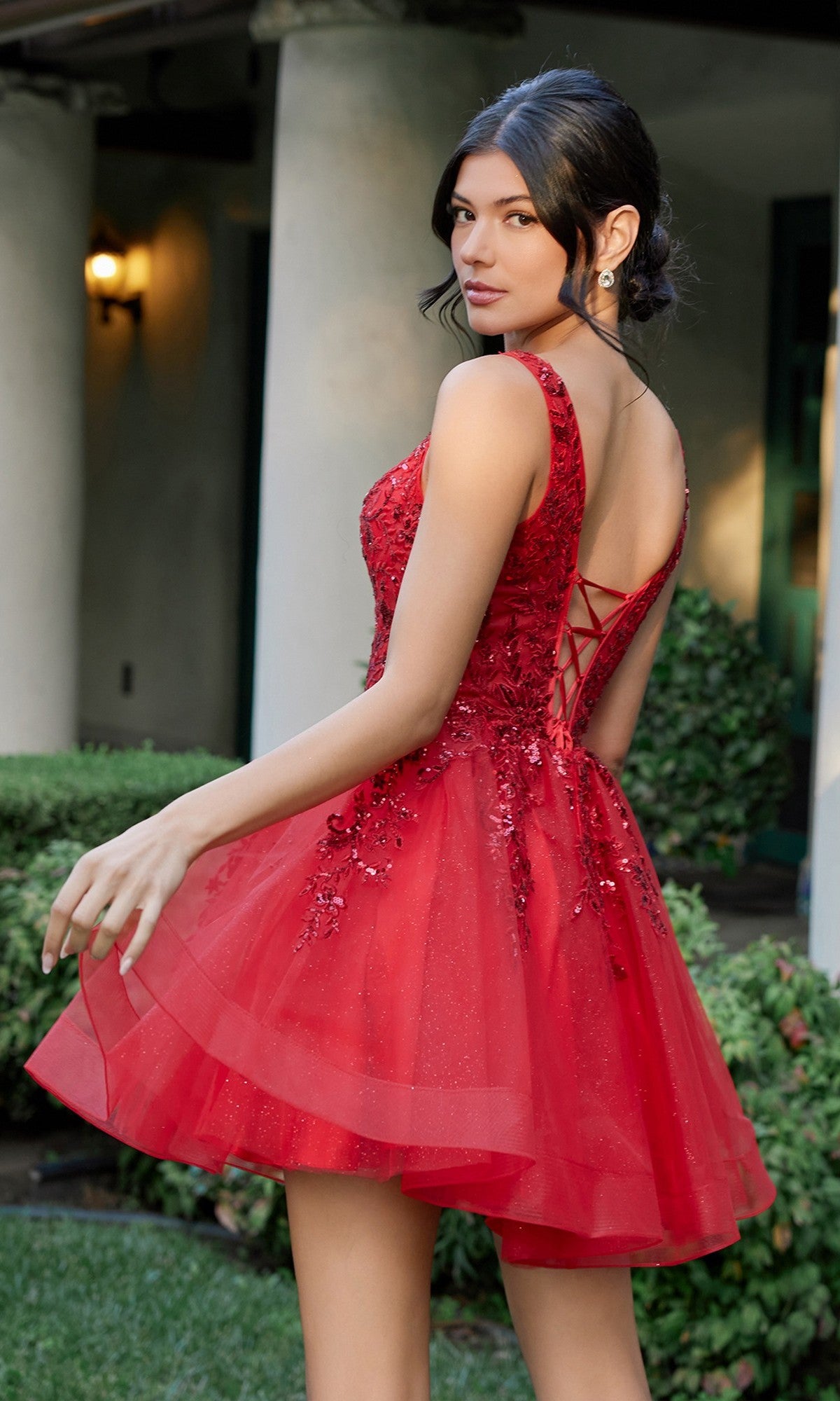 A woman stands outdoors in front of shrubs and a columned building, looking over her shoulder while wearing the Short Homecoming Dress: Juliet JT987N—a red, sleeveless dress featuring a lace-up back and an embroidered lace bodice.